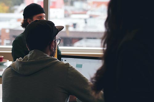 Three people are gathered indoors near a window; one in the foreground works on a laptop, perhaps researching The 9 Strongest Ecommerce SEO Tips for 2026, while two others, one blurred and one wearing a hat, chat. City buildings are visible outside. - Clio Websites | Top Canadian Web Design Company