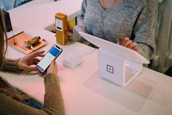 Two people at a counter; one pays with a smartphone using contactless payment while the other operates a white Square point-of-sale device. For businesses, mastering The 9 Strongest Ecommerce SEO Tips for 2026 can enhance visibility and sales. - Clio Websites | Top Canadian Web Design Company
