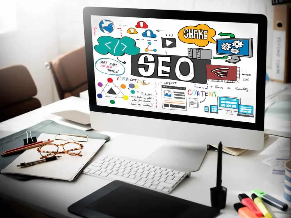 A computer monitor on a desk displays a vibrant digital collage with the words "Calgary SEO" in bold. The desktop includes notebooks, a pen, and a tablet. The room features chairs adorned with cushions. - Clio Websites | Top Canadian Web Design Company