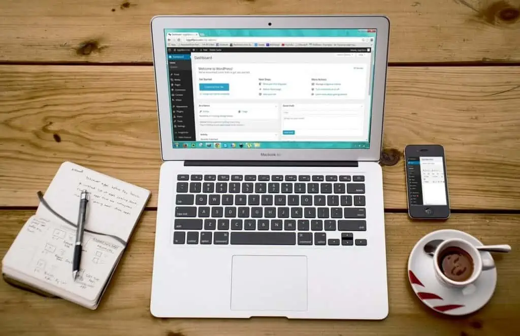 wordpress-developer-calgary A laptop showcasing a WordPress development dashboard is on a wooden table, flanked by a notebook filled with sketches and a smartphone. A cup of coffee sits nearby, and a pen is placed on the open notebook. - Clio Websites | Top Canadian Web Design Company