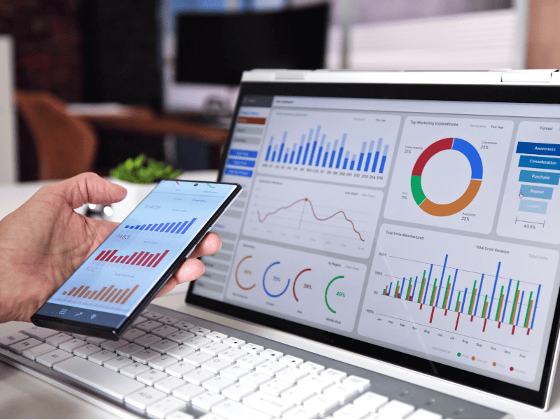 A person holds a smartphone displaying bar graphs next to a laptop featuring dashboards with bar charts, line graphs, and a pie chart, illustrating calgary seo data analysis or business analytics. - Clio Websites | Top Canadian Web Design Company Hire a WordPress Developer in the USA