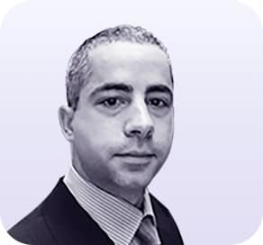 A man with short, dark hair wears a suit, dress shirt, and tie. He faces forward with a neutral expression. The light gradient background with rounded corners suggests a modern style often seen on about Clio websites. - Clio Websites | Top Canadian Web Design Company