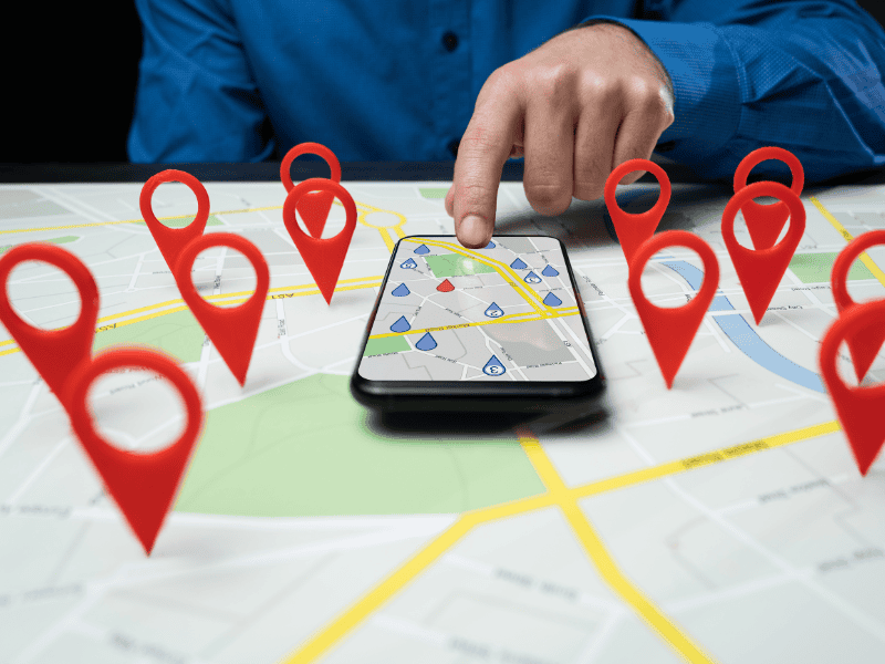 A person in a blue shirt points at a smartphone displaying a map, placed on a paper map with several red location markers—perfect for visualizing calgary seo strategies and local business targeting. - Clio Websites | Top Canadian Web Design Company