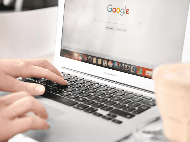 A person types on a MacBook Air laptop with the Google search homepage open, possibly researching Calgary SEO strategies. A cup, likely containing a beverage, sits in the foreground. - Clio Websites | Top Canadian Web Design Company