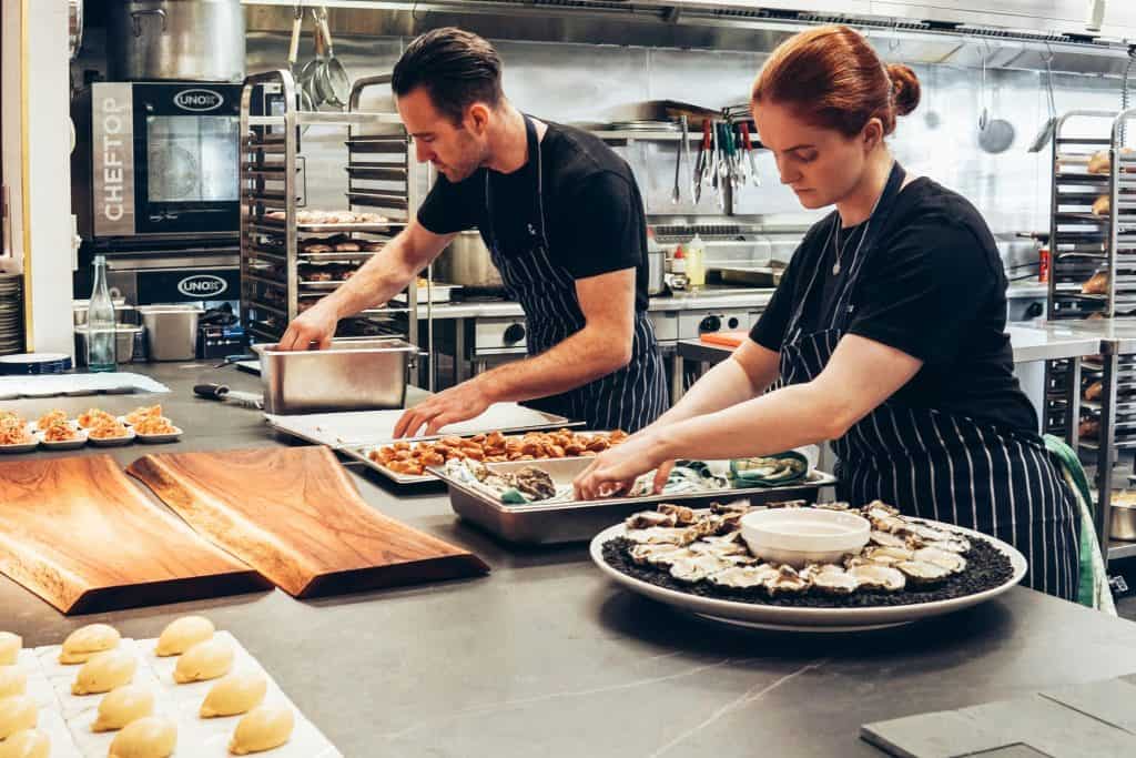 Two chefs in black shirts and striped aprons prepare food in a professional kitchen, arranging ingredients on trays and plates—much like small businesses organizing effective SEO strategies for success. A variety of dishes and utensils are spread across the countertop. - Clio Websites | Top Canadian Web Design Company