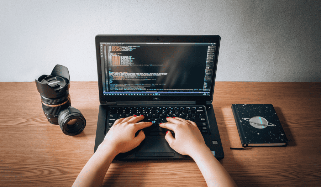 A person typing on a laptop displaying lines of code, with a camera lens, camera body, and a notebook with a planet illustration placed on a wooden desk—perfect for showcasing managed website services in action. - Clio Websites | Top Canadian Web Design Company
