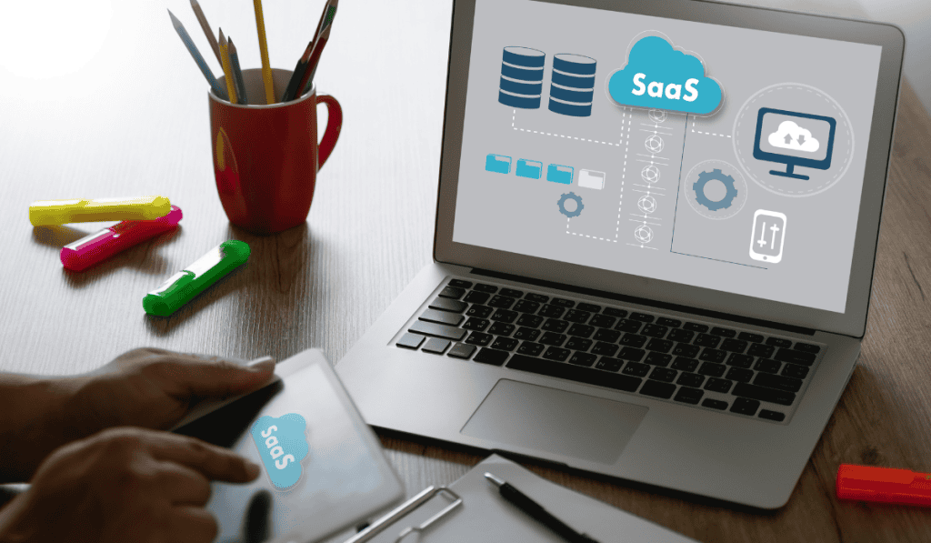 A person uses a tablet and a laptop at a desk with office supplies. The laptop screen displays a cloud SaaS graphic with icons for servers, data storage, workflow diagrams, and highlights of the best WooCommerce options. - Clio Websites | Top Canadian Web Design Company