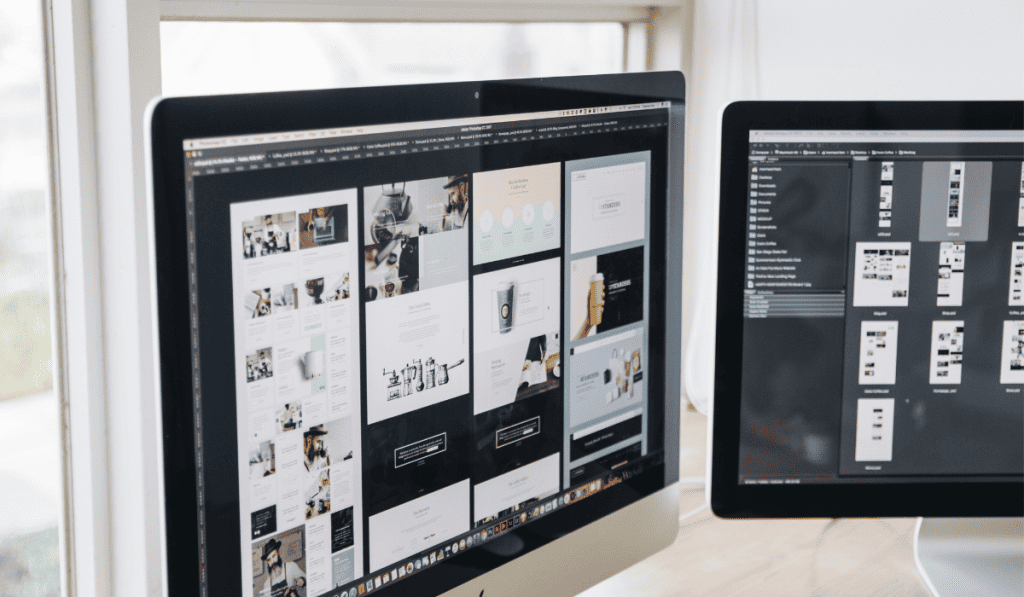 Two computer monitors on a desk display web design layouts and WooCommerce Comparison charts. The workspace is next to a window, with various website pages and design elements visible on the screens. - Clio Websites | Top Canadian Web Design Company