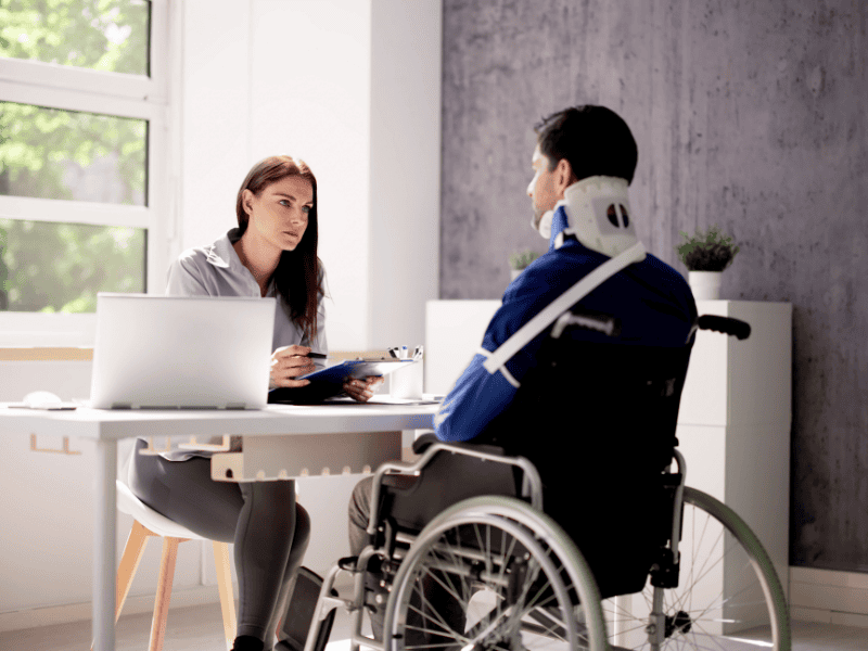 A woman sits at a desk with a laptop and clipboard, attentively speaking to a man in a wheelchair who has a neck brace and arm sling, in a bright office designed for a personal injury lawyer website. - Clio Websites | Top Canadian Web Design Company