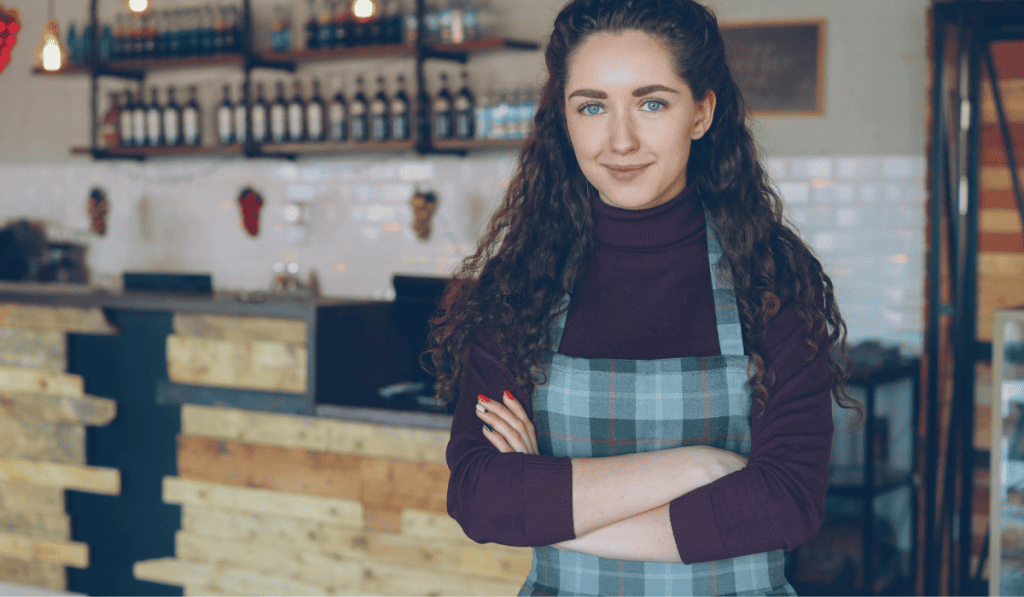 A young woman with curly brown hair, wearing a plaid apron and maroon turtleneck, stands confidently with arms crossed in a rustic café—perfectly reflecting the dedication you'd expect from a WordPress website speed optimization Calgary expert. - Clio Websites | Top Canadian Web Design Company