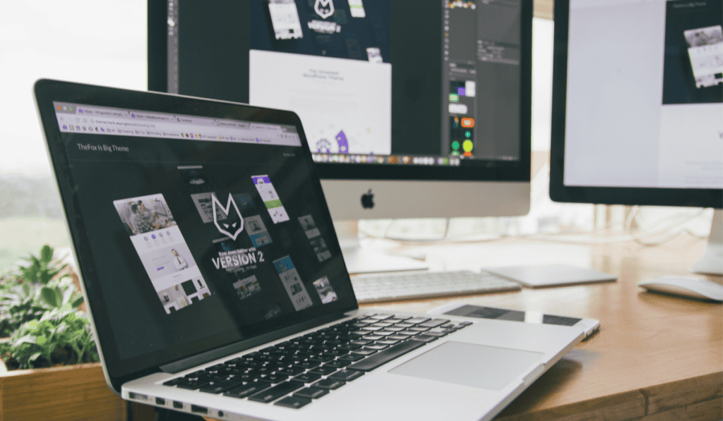 A laptop and two desktop monitors on a wooden desk display website design work. The screens showcase a Wordpress website for small business Calgary, featuring “Version 2,” webpage layouts, and a design application. - Clio Websites | Top Canadian Web Design Company