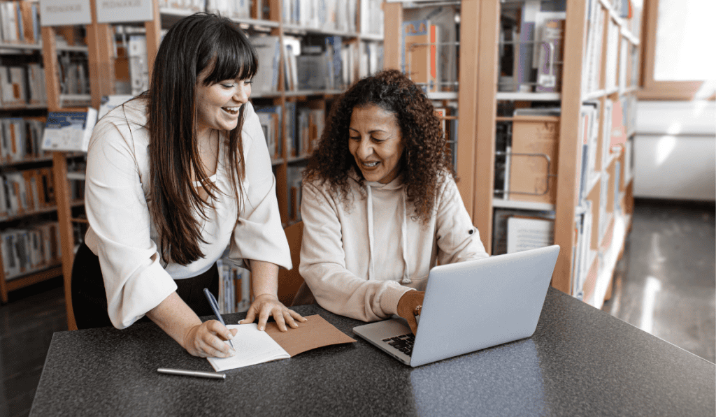 Two women in a library smile while working together at a table—one writes in a notebook, the other uses her laptop to research wordpress website for nonprofits Calgary, with bookshelves in the background. - Clio Websites | Top Canadian Web Design Company