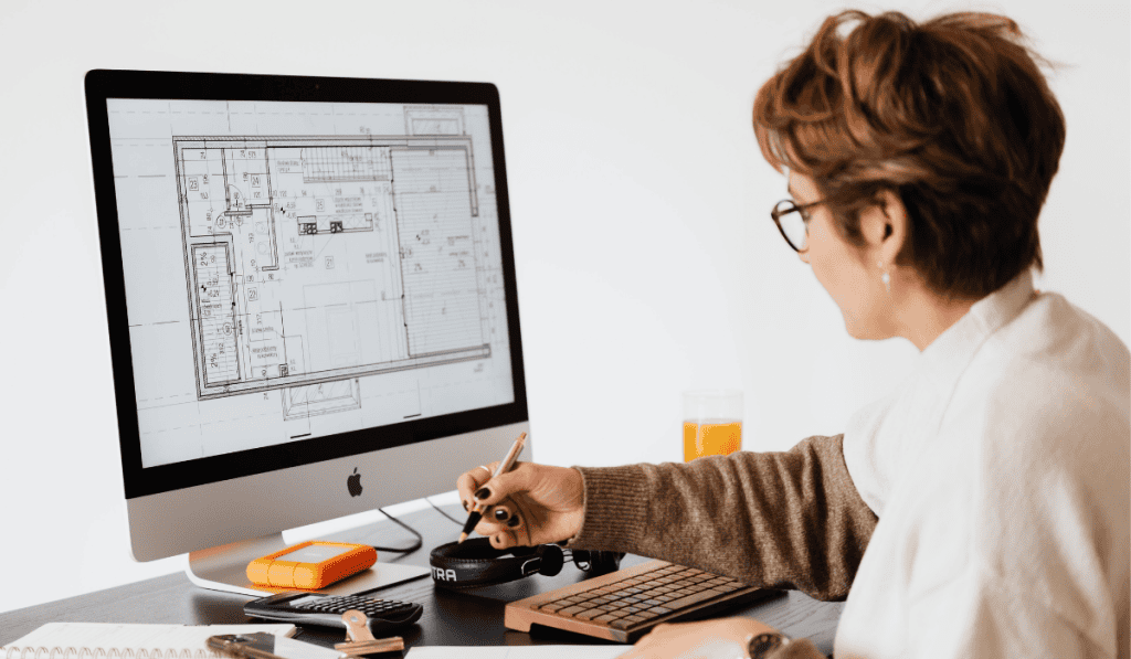 A person wearing glasses is sitting at a desk, working on architectural blueprints for a WordPress website for nonprofits Calgary, displayed on a desktop computer screen. The workspace includes a keyboard, external drives, notepad, and a glass of orange juice. - Clio Websites | Top Canadian Web Design Company