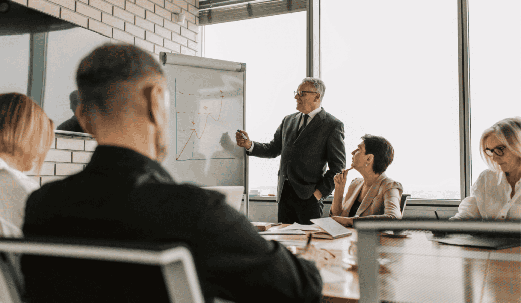 A man in a suit presents a chart on a flipboard to four colleagues seated around a conference table in a modern office, discussing web management services tailored for small businesses. - Clio Websites | Top Canadian Web Design Company