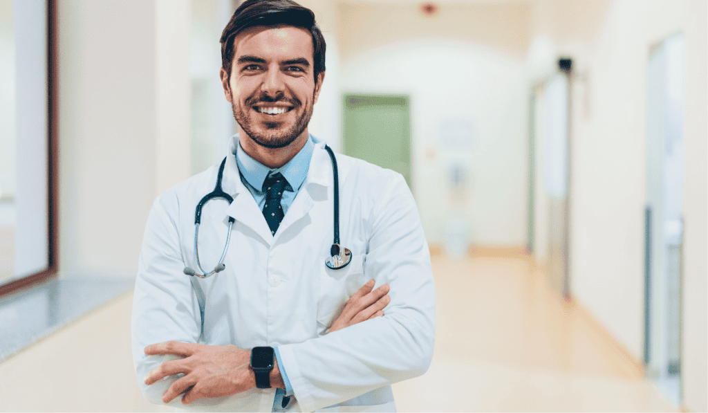 A smiling male doctor with dark hair and a beard stands in a hallway, wearing a white coat, stethoscope, blue shirt, and tie, with arms crossed—a professional image perfect for showcasing Calgary website design for doctors. - Clio Websites | Top Canadian Web Design Company