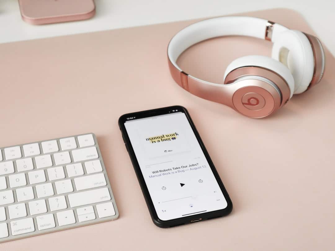 A smartphone playing a podcast rests on a pink desk next to white headphones, a white keyboard, and a closed laptop, all on a pink surface. - Clio Websites | Top Canadian Web Design Company