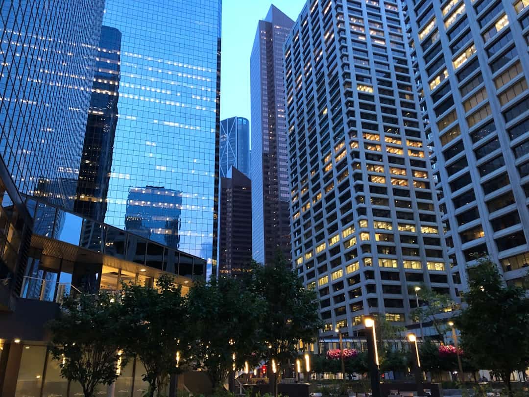 Cityscape at dusk with tall glass and concrete office buildings, illuminated windows, green trees, and streetlights glowing in the foreground. - Clio Websites | Top Canadian Web Design Company